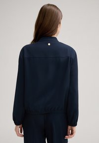 Marineblaue Jacke mit klassischem Kragen, elastischen Bündchen und einem goldfarbenen Akzentknopf auf der Rückseite. Hergestellt aus glattem, leichtem Material.