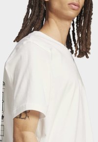 Balta kokvilnas īsroku t-krekls ar apaļu kakla izgriezumu un grafiskiem attēliem vienā pusē. Gluda tekstūra, standarta piegriezums.