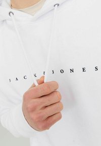 Hoodie blanc en coton, avec une capuche à cordon et un logo frontal "JACK & JONES" en noir. Texture douce, design décontracté.