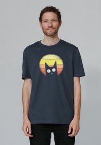 T-shirt en coton bleu foncé avec un graphisme d'un chat noir sur un design circulaire de coucher de soleil dans des tons jaunes, oranges et rouges.