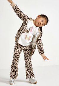 Veste et pantalon assortis à imprimé léopard, avec une chemise blanche ornée de motifs colorés de papillons. Des baskets argentées métalliques complètent la tenue.