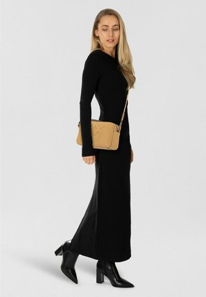 Lange zwarte jurk met een nauwsluitend ontwerp en lange mouwen, gecombineerd met een zandkleurige crossbodytas met reliëfdetails en zwarte enkellaarzen met hak.