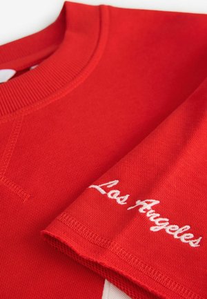 Sudadera roja con cuello acanalado y "Los Ángeles" bordado en blanco en la manga. Tejido texturizado con un diseño de ajuste relajado.