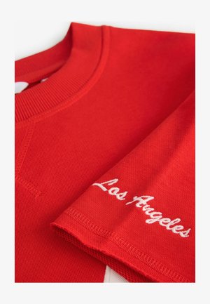 Raudonas džemperis su raukiniais apykakle ir balta siuvinėta "Los Angeles" ant rankovės. Tekstūruotas audinys su laisvu kirpimu.