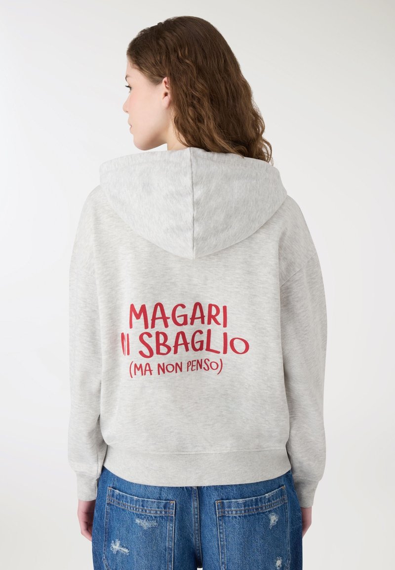 Giovane donna con capelli ricci che indossa una felpa con cappuccio grigio chiaro con testo italiano rosso sulla schiena e jeans blu strappati, vista di spalle.