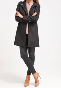 Sort uldfrakke med hætte, to knapper foran, og en let skræddersyet pasform. Bæres med mørke jeans og nude højhælede sko.