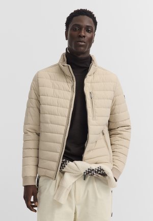 Young man wearing a beige quilted jacket over a black turtleneck sweater and light-colored pants with a patterned sweater tied at the waist.