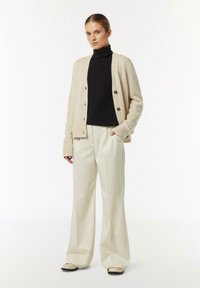 Beżowy kardigan z dzianiny z zapięciem na guziki, noszony na czarnym golfie, w zestawie z kremowymi spodniami typu wide-leg i czarnymi butami, o gładkiej teksturze.