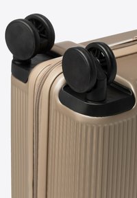 Close-up van de hoek van een beige harde koffer met twee zwarte, dubbele spinners wielen en een ritsnaad.