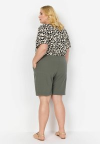Blus med kort ärm i svart och krämfärgad mönster, kombinerad med olivgröna shorts. Shortsen har sidofickor och en avslappnad passform. Sandaler bärs.