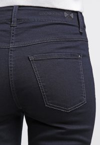 Mørke denimjeans med baklomme-design og subtil stikning. Livremen har brandingdetaljer og en teksturert lapp.