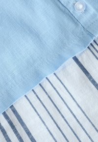 Himmelblauem Stoff mit einem Rautenmuster, genäht an einem weißen Stoff mit dünnen blauen Streifen. Enthält ein Detail aus Kunststoffknopf.