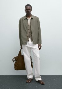 Oliven grøn skjorteblusejakke, beige cardigan, hvide bukser, brun ruskinds taske og sorte sandaler på en mørk overflade med en lys baggrund.