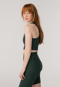 Ung kvinne med langt rødt hår som har på seg en mørkegrønn ermeløs crop-top og høytlivs sykkelshorts, står mot en ensfarget bakgrunn.