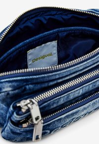 Denim-Mäppchen mit navy-farbenem Innenfutter, ausgestattet mit zwei Reißverschlussfächern, silbernem Zubehör und einem Etikett mit der Aufschrift "Desigual". Strukturierte Oberfläche und Nähte.
