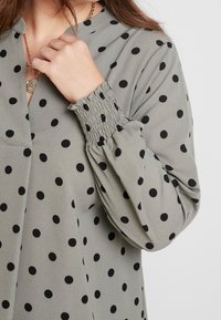 Blouse verte à pois noirs, avec un col boutonné, des manches froncées et une texture fluide pour le confort.