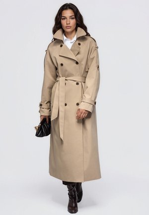Une femme porte un long trench beige ceinturé, une chemise blanche et des bottes noires. Elle tient un petit sac à main noir sur un fond de studio uni.