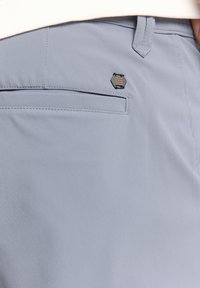 Lichtblauwe broek van gladde stof, met een achterzak en een klein zwart en gouden logootje vlakbij de rand van de zak.