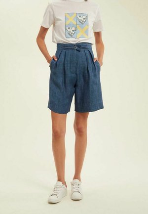 Shorts en denim taille haute avec des plis, dotés de poches latérales ; associés à un t-shirt blanc affichant un design graphique bleu et jaune.