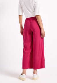 Rosa vida byxorna med avslappnad passform, texturerad tyg, och hög midja. Modell som bär vita skor, beskuren vy bakifrån.