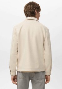 Uomo con capelli castani mossi che indossa una giacca color crema e jeans grigi, visto di spalle su uno sfondo bianco semplice.