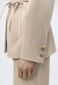 Μπεζ σακάκι κομμένο και ραμμένο, με σφηκοφωλιά στη μέση, υφή υφάσματος και δύο μεγάλα κουμπιά στο μανίκι. Διαθέτει πλαϊνή τσέπη.