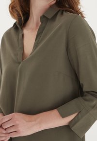 Blouse vert olive en tissu lisse, avec un col doux, un décolleté en V et des manches trois-quarts avec des poignets retroussés. Design simple, sans motifs.