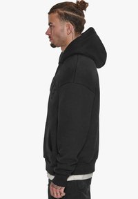 Sort hoodie lavet af tykt stof, med en stor frontlomme og justerbar hætte. Ærmerne er løstsiddende og ribbet ved manchetterne.