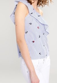 Gestreiftes blau-weißes ärmelloses Oberteil mit Rüschenausschnitt und gestickten floralen Akzenten; getragen mit weißen Hosen; lässiger Stil.