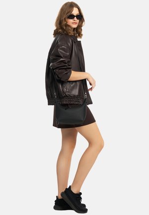 Kvinde iført mørk læderjakke, solbriller, sort mininederdel, sorte sneakers og bærende på en sort skuldertaske, stående i sidepose.