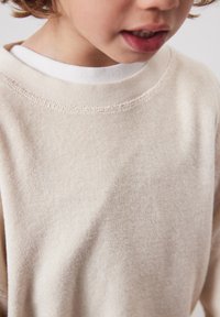 Enfant portant un pull à manches longues beige texturé sur un t-shirt blanc à col rond, lèvres légèrement entrouvertes, sur un fond uni.