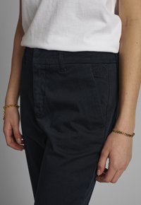 Pantalon en coton noir à coupe ajustée, doté de poches latérales et d'un détail de logo subtil sur la ceinture, associé à une chemise blanche.