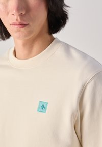 Beige sweatshirt met een ronde hals, voorzien van een klein turquoise geborduurd logo op de borst. Zachte, gladde textuur.