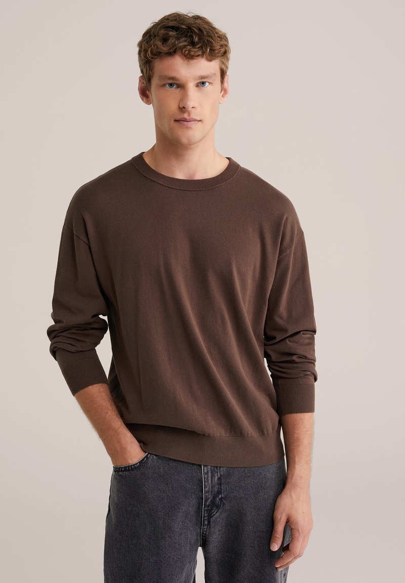 Pull en maille marron avec un col rond et des manches longues. Présente des poignets et un ourlet côtelés, associé à un pantalon gris foncé. Texture lisse.