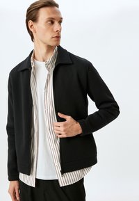 Chaqueta negra con cremallera y cuello puntiagudo, llevada sobre una camisa blanca con rayas rojas, superpuesta con una camiseta blanca debajo.