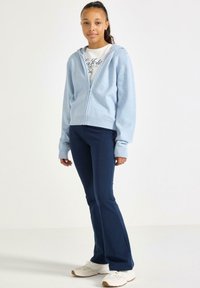 Jasnoniebieska bluza z zamkiem błyskawicznym, z ribbowanym dołem i mankietami, w zestawie z granatowymi spodniami w typie dzwonów i białymi butami sportowymi. Szczegółowy grafik na koszulce.