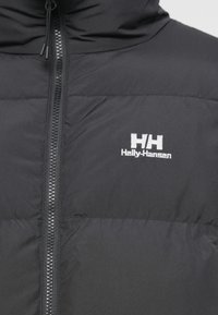 Schwarze isolierte Jacke mit hohem Kragen, ausgestattet mit einem Reißverschluss vorne und dem weißen Helly Hansen-Logo im Brustbereich. Glatte Textur.