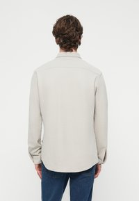 Világosszürke ing texturált anyagból, gombos gallérral, hosszú ujjakkal és ívelt szegéllyel. A hátulnézet finom varrási részleteket mutat.