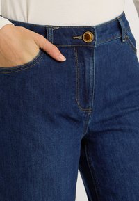 Primer plano de una persona con jeans azul oscuro, con una mano descansando dentro del bolsillo frontal y un botón dorado visible en la cintura.