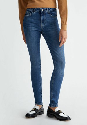 Frau trägt enge blaue Jeans, braunes langärmeliges Oberteil und schwarz-weiße Loafer und steht vor einem einfarbigen Hintergrund.