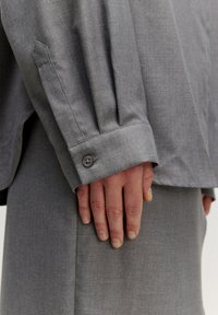 Graues Hemd mit strukturiertem Kragen, umgeschlagenen Ärmeln, subtiler Textur und einer Nahaufnahme einer Hand, die auf dem Stoff ruht.