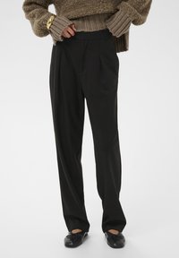 Crne krojene hlače s plisiranim detaljima, visokog struka, od laganog materijala, uparene s bež teksturiranom pletenom svjetlošću i crnim baletankama.