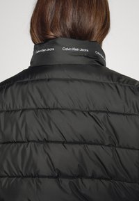 Giubbotto puffer nero con texture trapuntata. Presenta un colletto alto con il logo "Calvin Klein Jeans" stampato lungo il bordo.