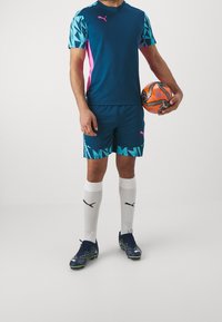 Sportovní oblečení Puma v námořnické modři s modrými a růžovými akcenty. Obsahuje tričko, šortky, bílé ponožky a černé kopačky se zelenými detaily. Drží fotbalový míč.
