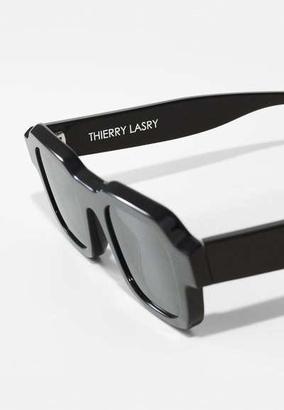 Mustad mustadriga prillid, millel on jäme ja nurkline disain. Tumedad läätsed ja sile mattviimistlus. "THIERRY LASRY" on templile trükitud.