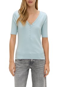 Lichtblauw geribd t-shirt met korte mouwen en een V-hals, voorzien van een knopenrij met vijf knopen. De stof is zacht en heeft een slim fit ontwerp. Gecombineerd met grijze jeans.