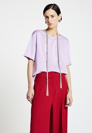 Top in raso lilla con maniche corte, abbinato a pantaloni larghi a vita alta rossi e una lunga collana di corda grigia con nappine.