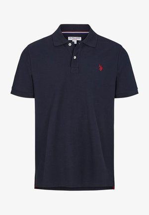 Man draagt een marineblauw poloshirt met korte mouwen en een rood geborduurd logo, gecombineerd met beige broek, staand tegen een effen achtergrond.