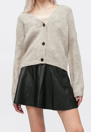 Femme portant un cardigan en maille beige à col en V avec des boutons noirs, associé à une mini-jupe plissée noire en simili cuir.
