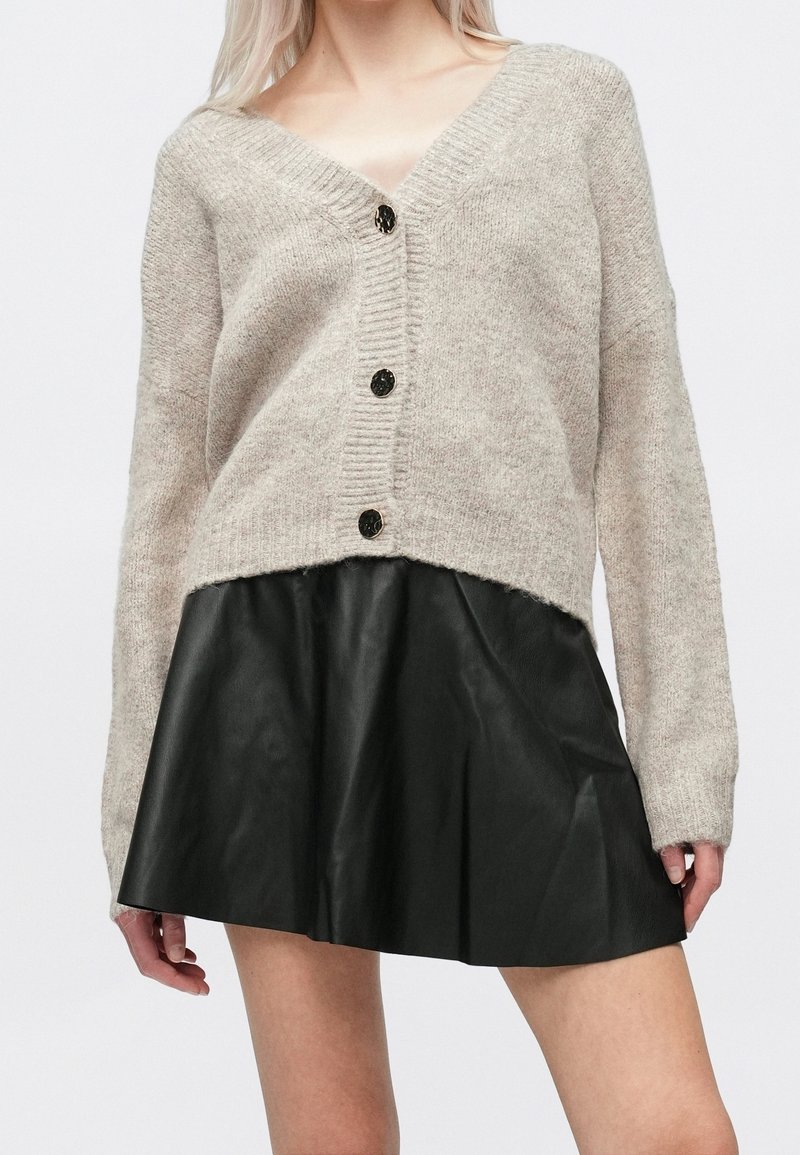 Femme portant un cardigan en maille beige à col en V avec des boutons noirs, associé à une mini-jupe plissée noire en simili cuir.